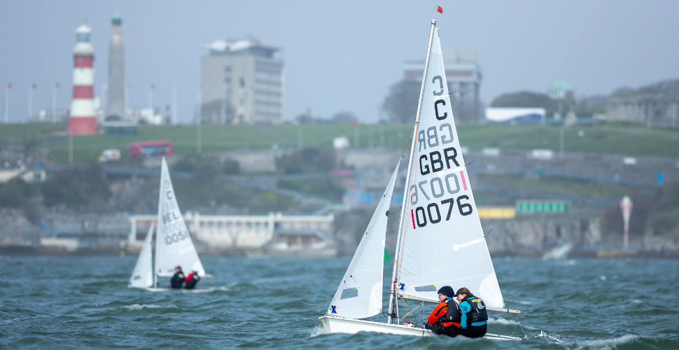 Sailors on a boat ont he water with Smeaton's Tower and Plymouth in the background